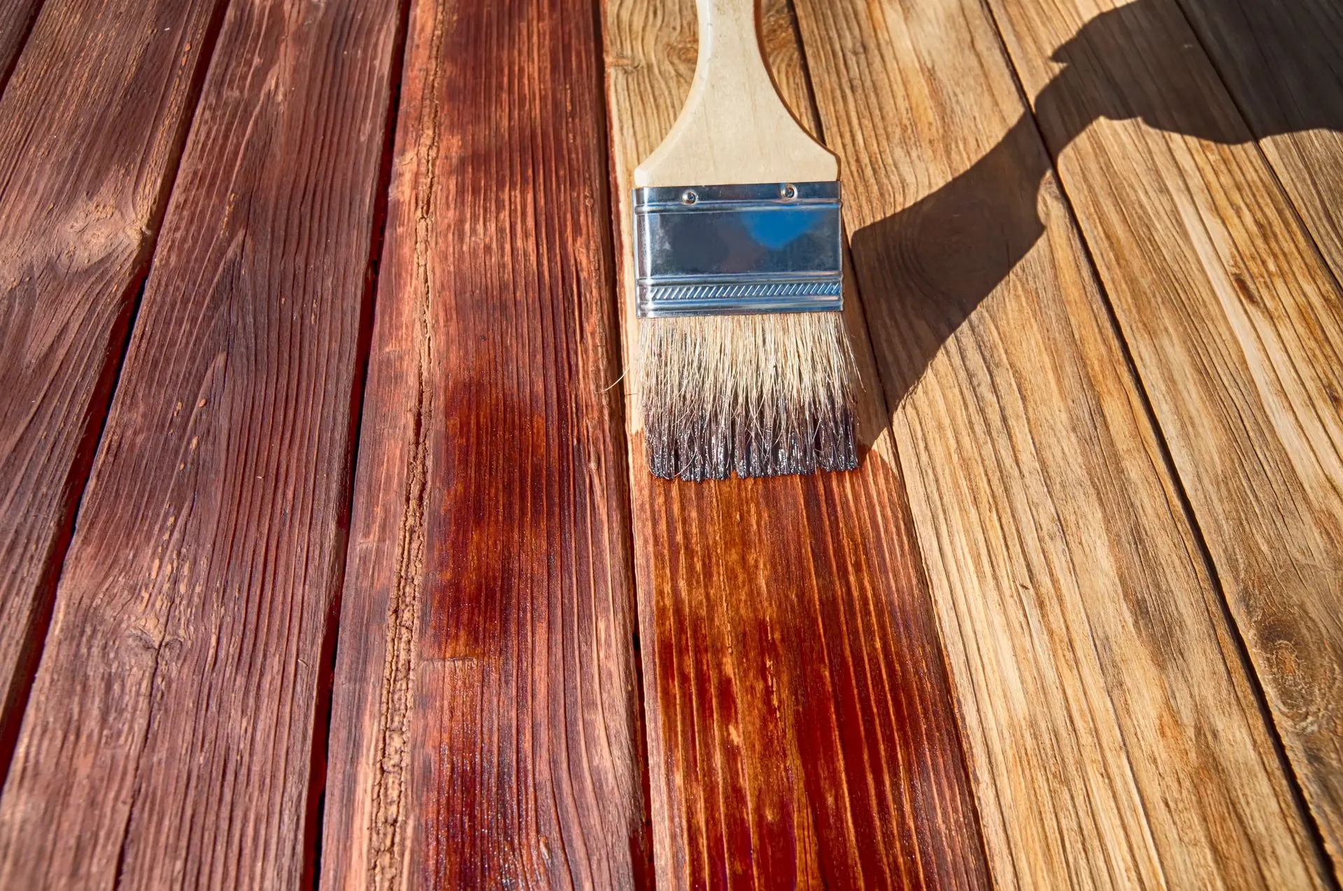 A paintbrush applying dark wood stain to deck boards, blending into the lighter untreated wood.