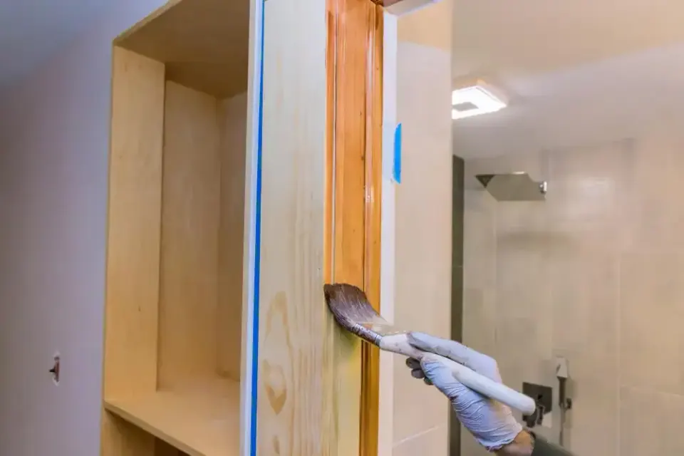 Close-up of a gloved hand brushing stain onto raw wood cabinet trim in a bathroom.