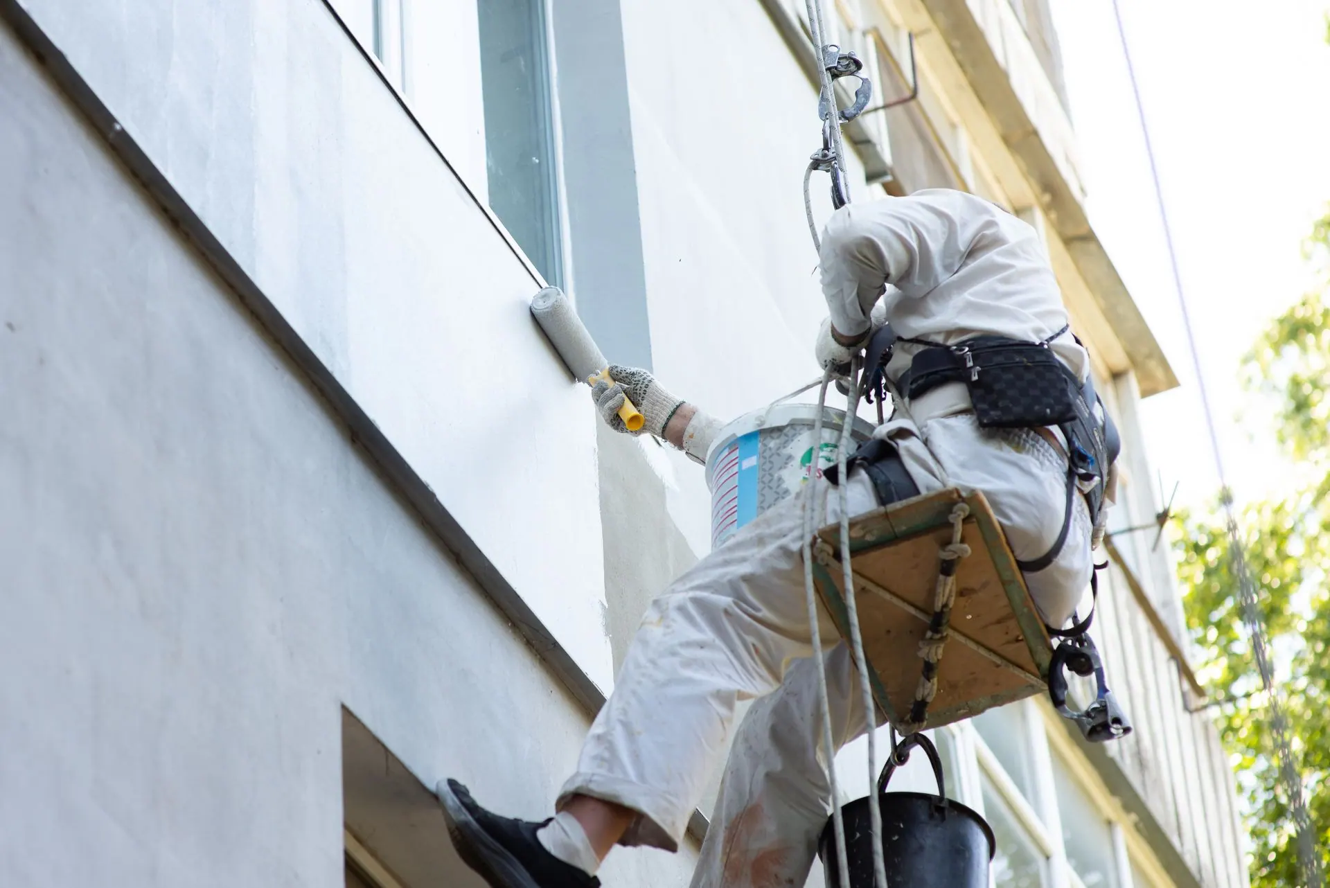 A painter suspended by rope and harness rolls paint onto a building's exterior wall.