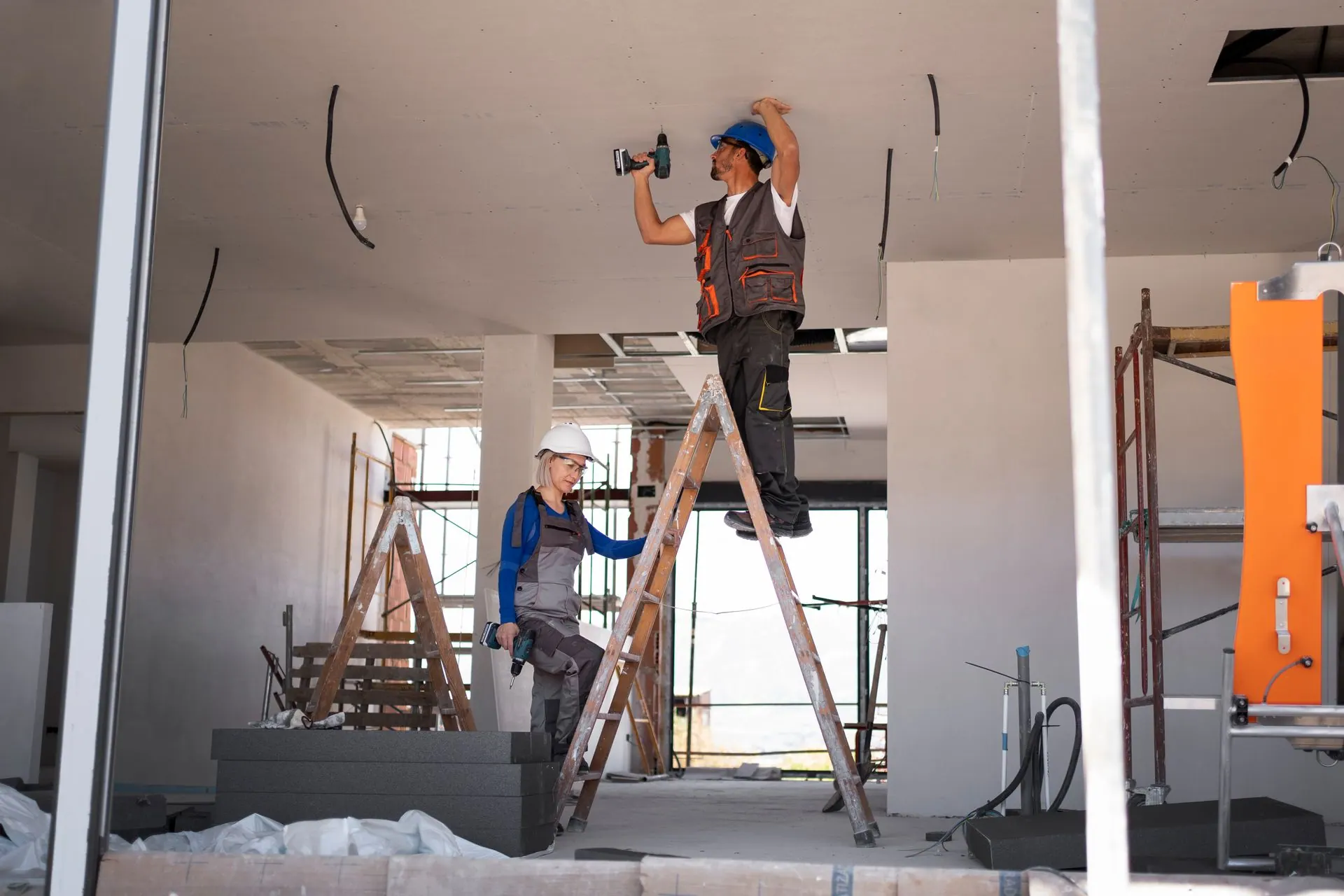 Two construction workers on a ladder installing drywall on a ceiling at a job site.