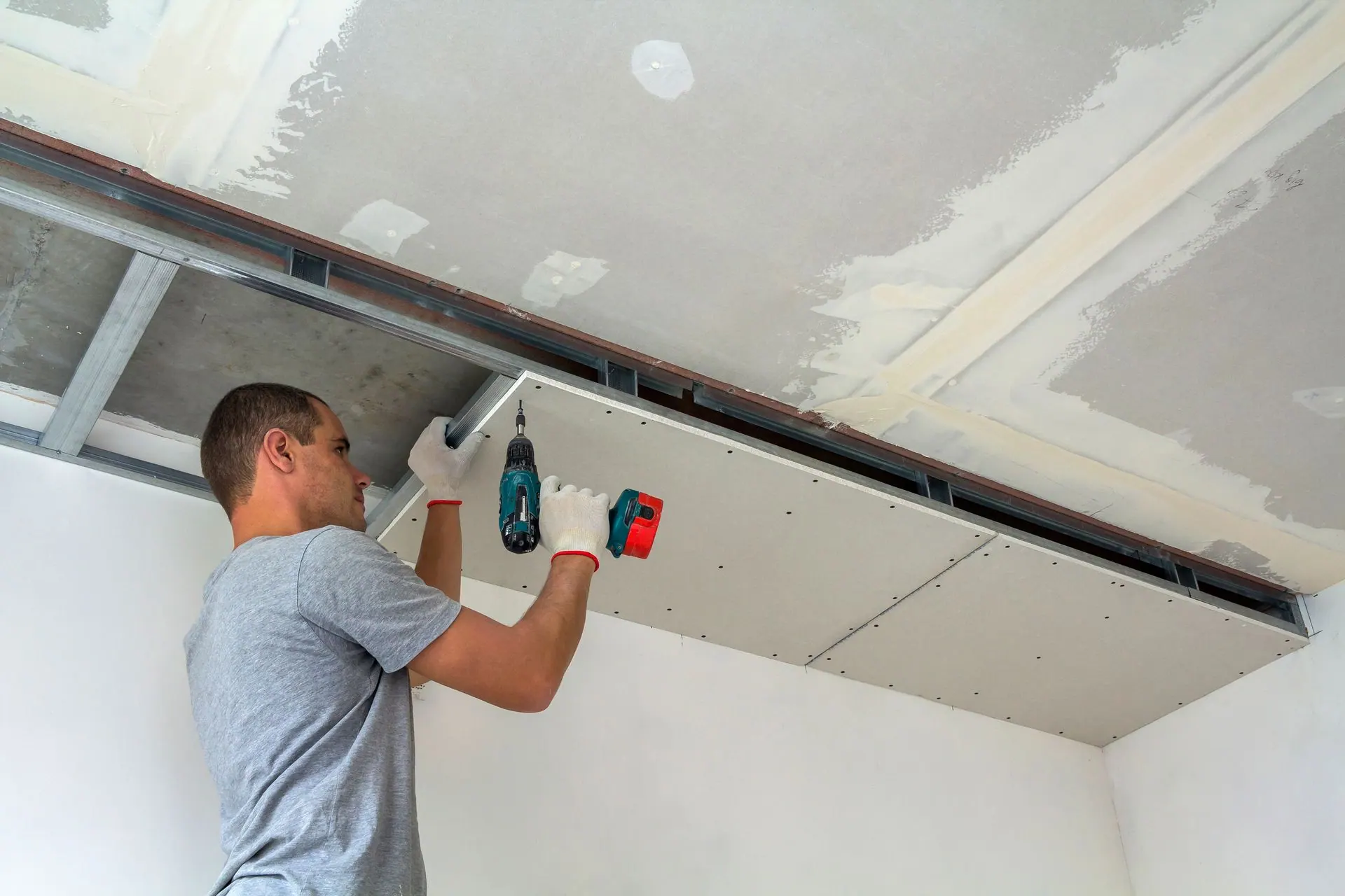 A worker uses a power drill to fasten a drywall panel to a ceiling frame.