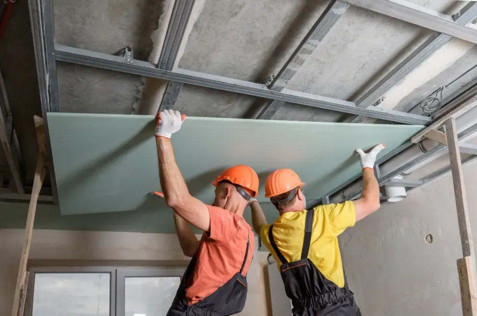 Two installers in hard hats securing a drywall panel to overhead ceiling framing in Catawba County.