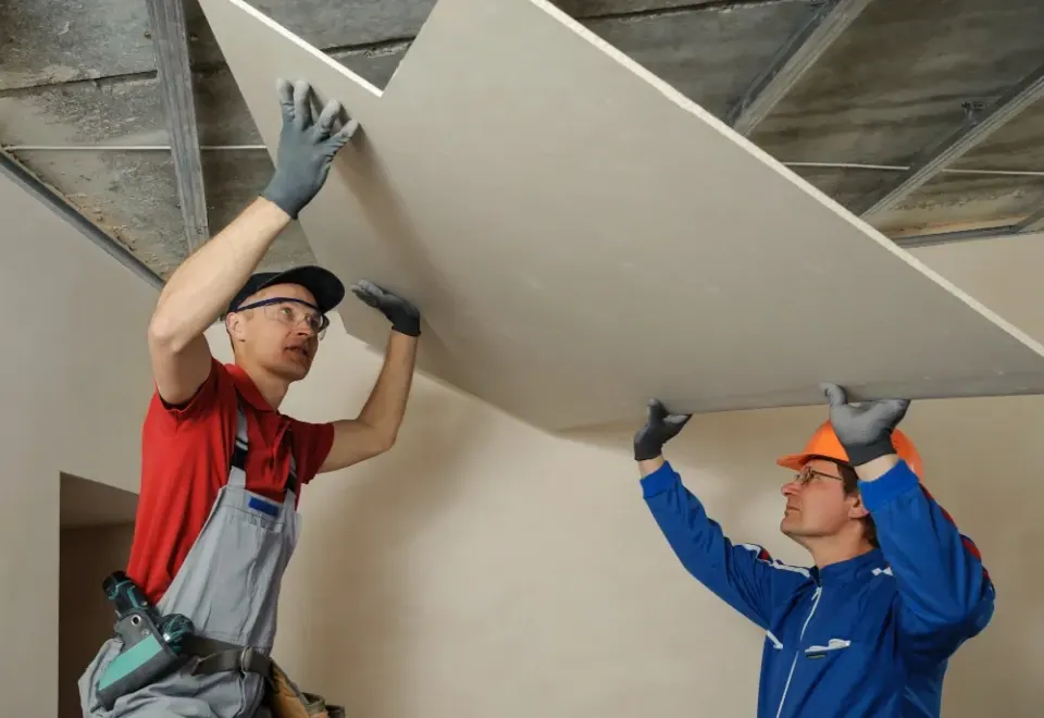 Two installers hoisting a large drywall sheet into position on an exposed ceiling frame.