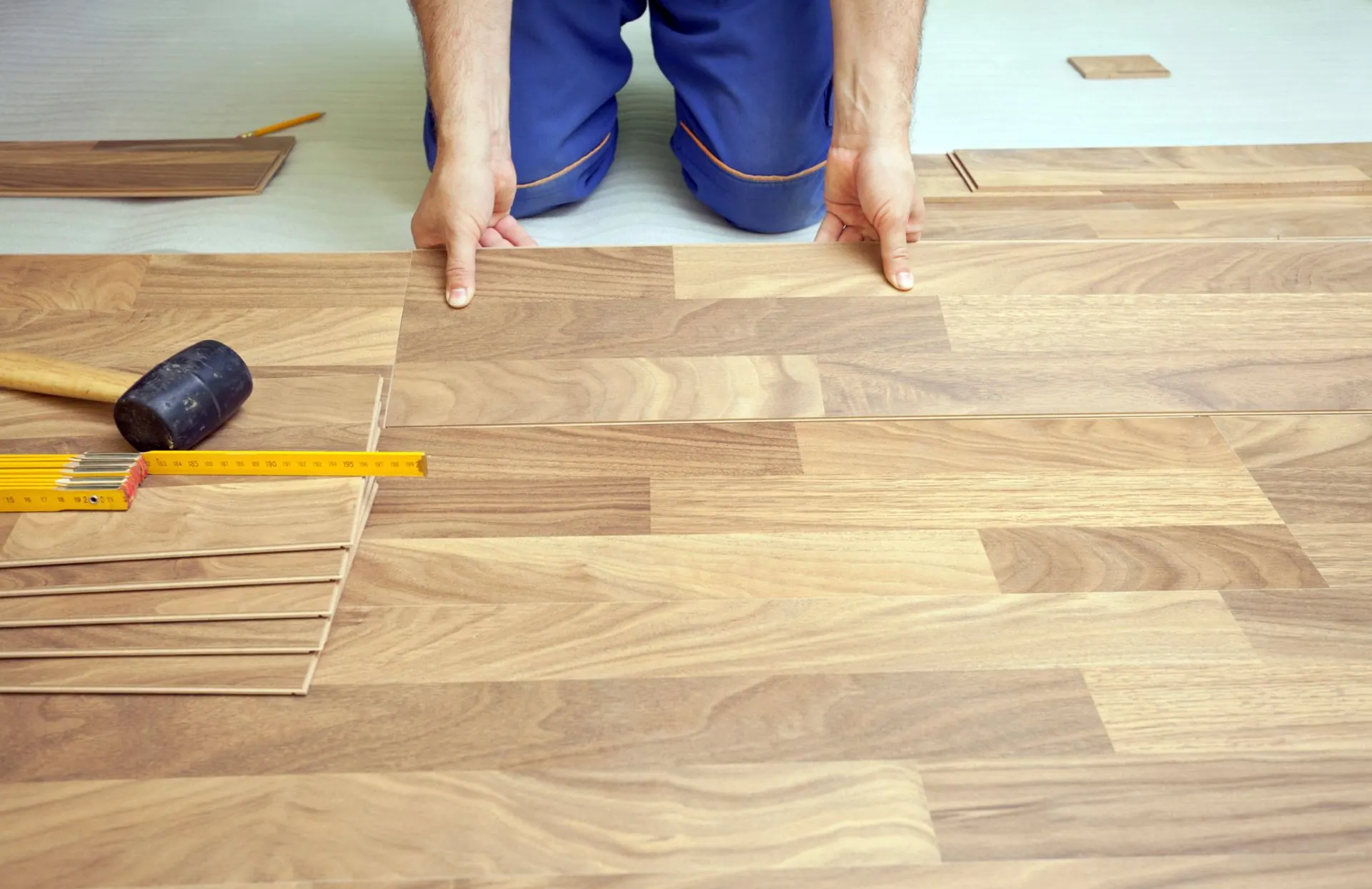 Installer kneeling to fit laminate wood-look flooring planks with a rubber mallet and tape measure nearby.