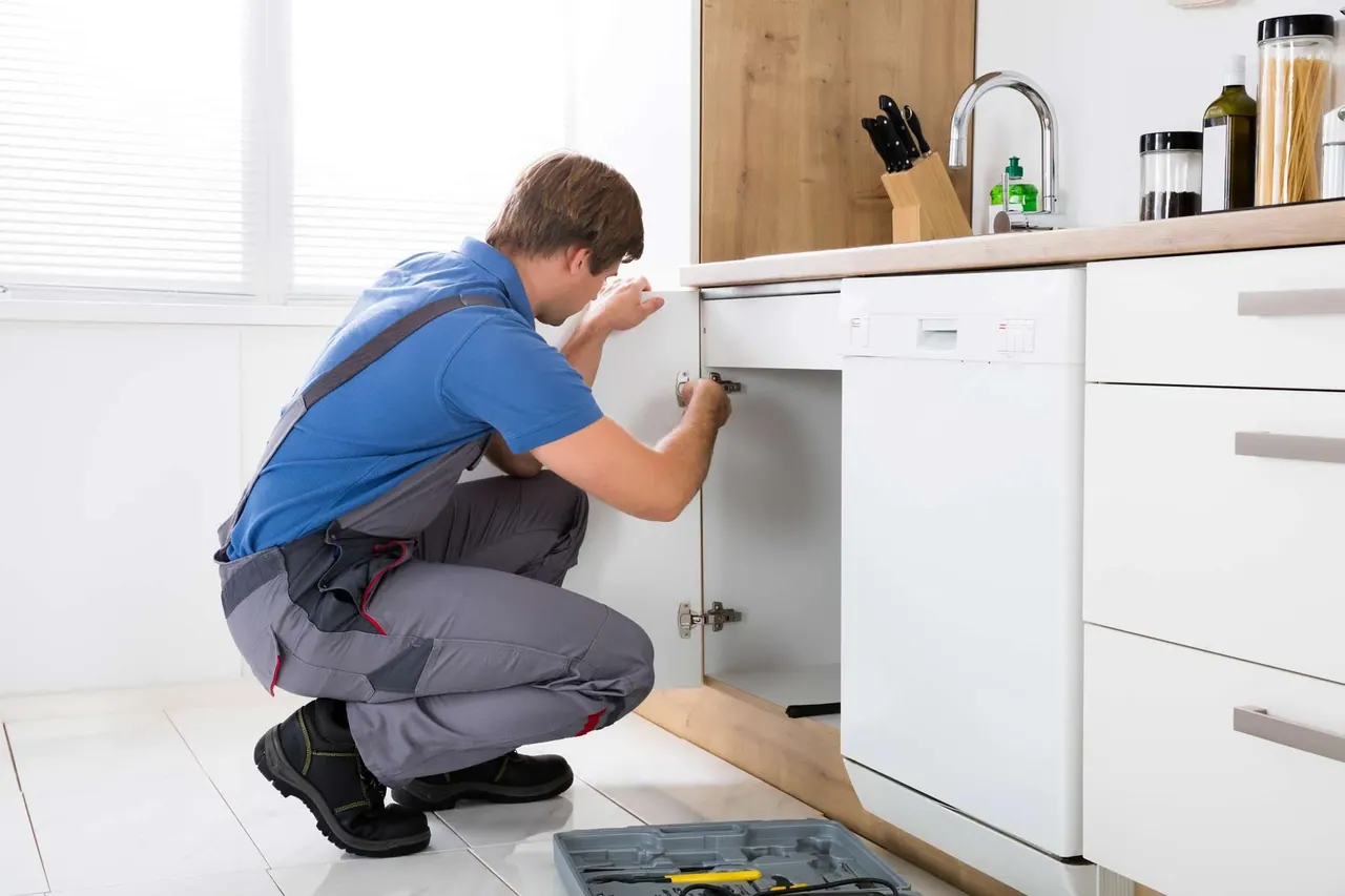 A technician adjusts a cabinet hinge under the counter in a white kitchen.