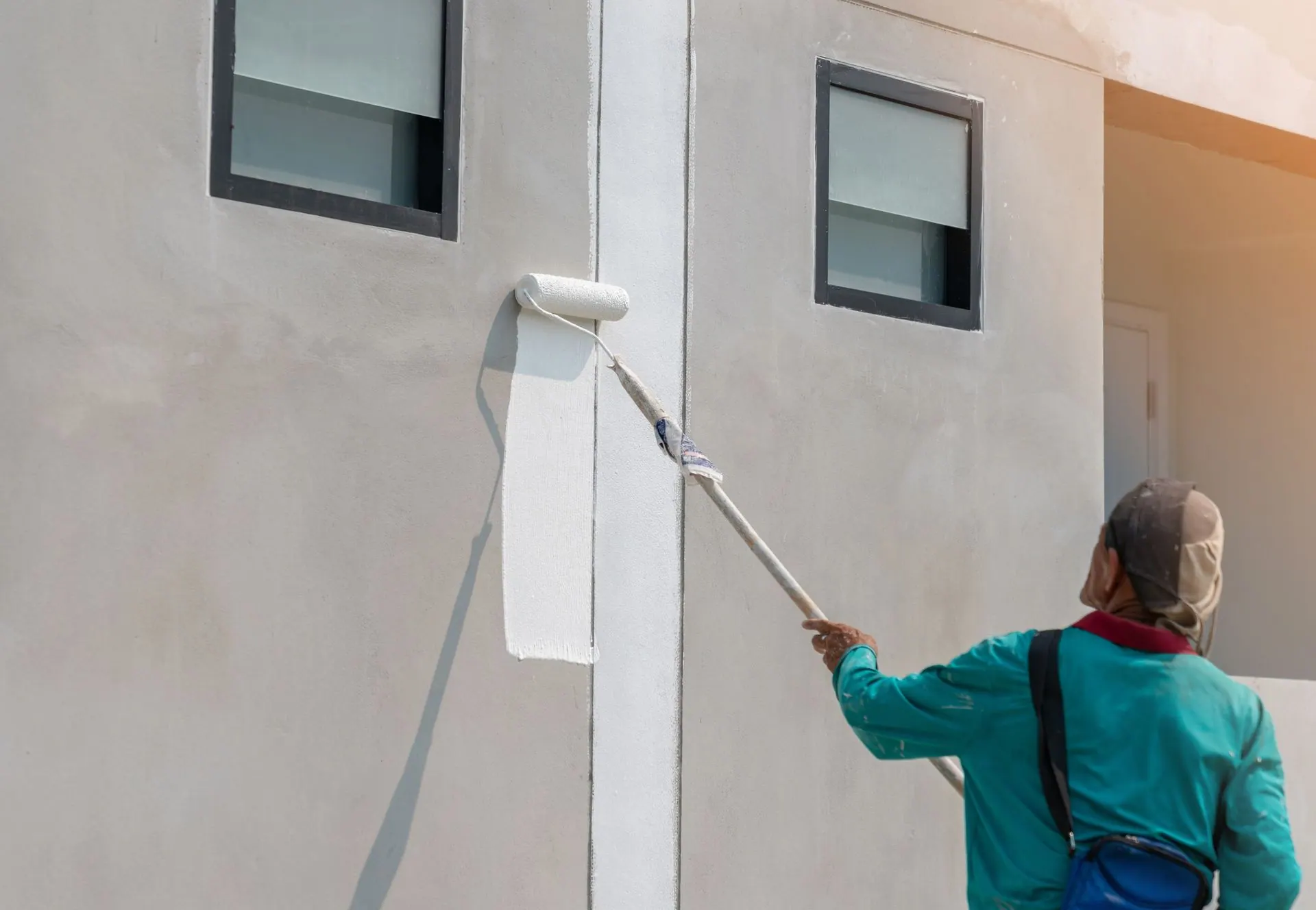 A painter uses an extension pole roller to paint a building exterior white.