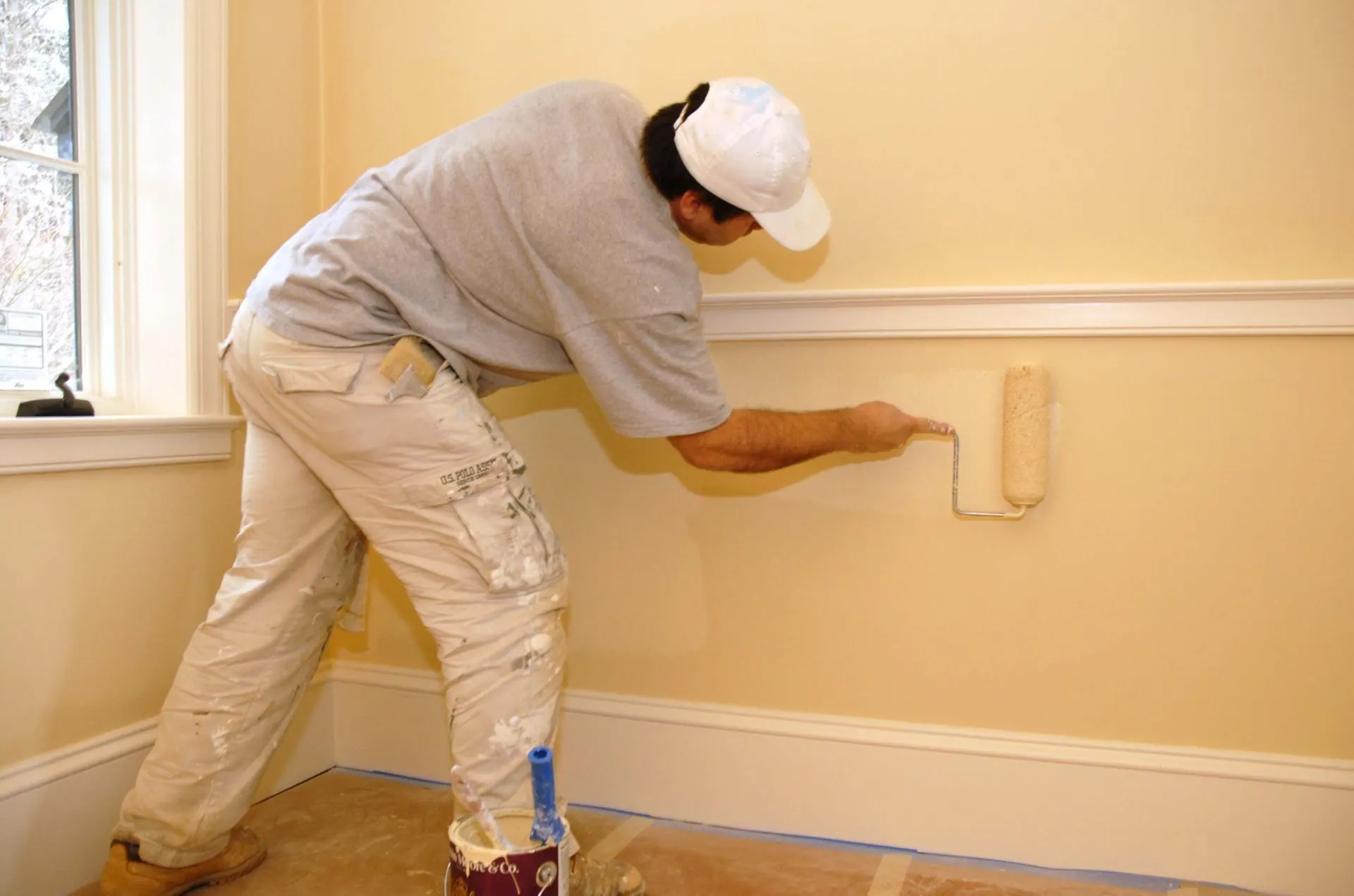 A painter rolling pale yellow paint onto a wall above the baseboard trim.