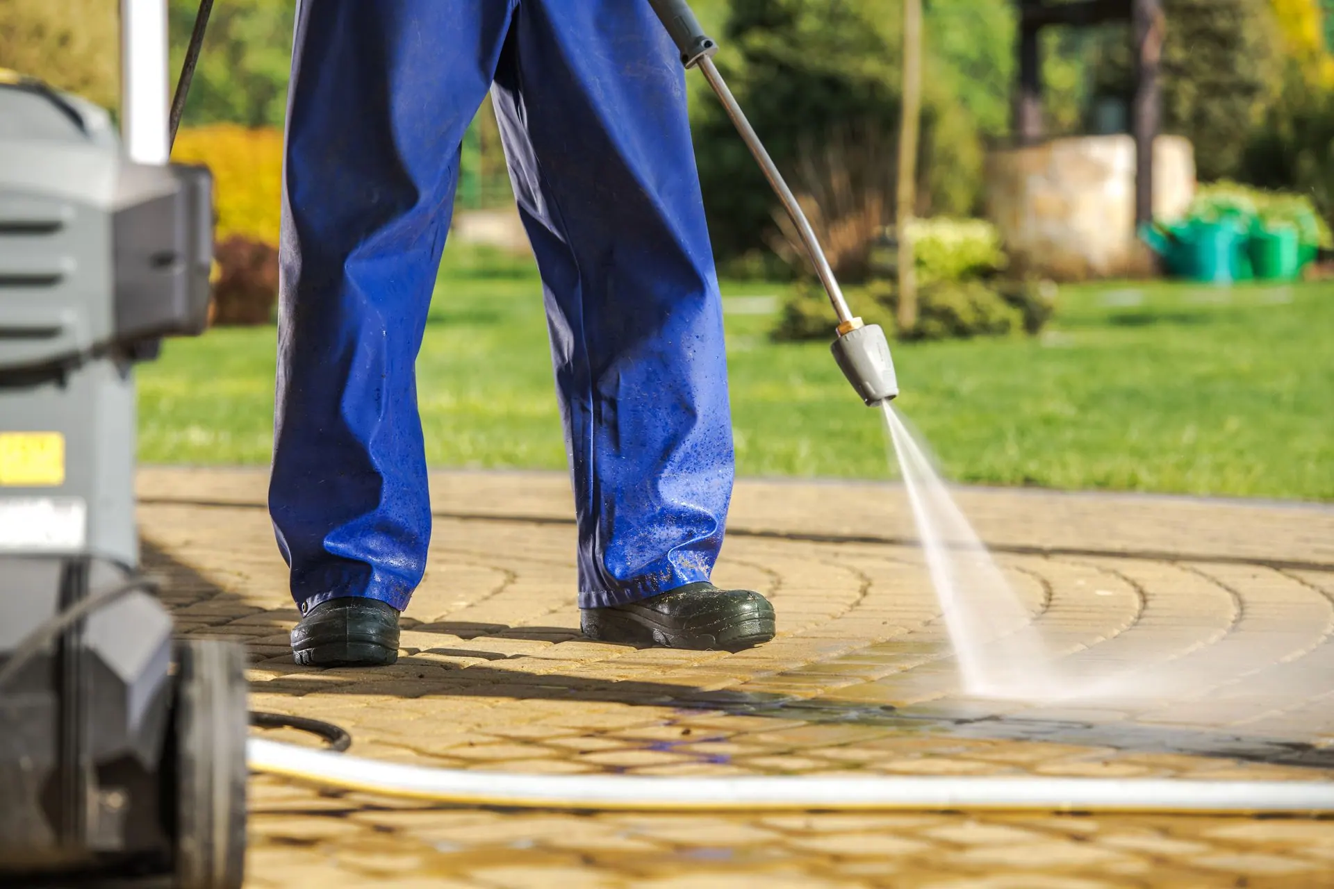 A worker in blue coveralls pressure washes a paver walkway beside a lawn.