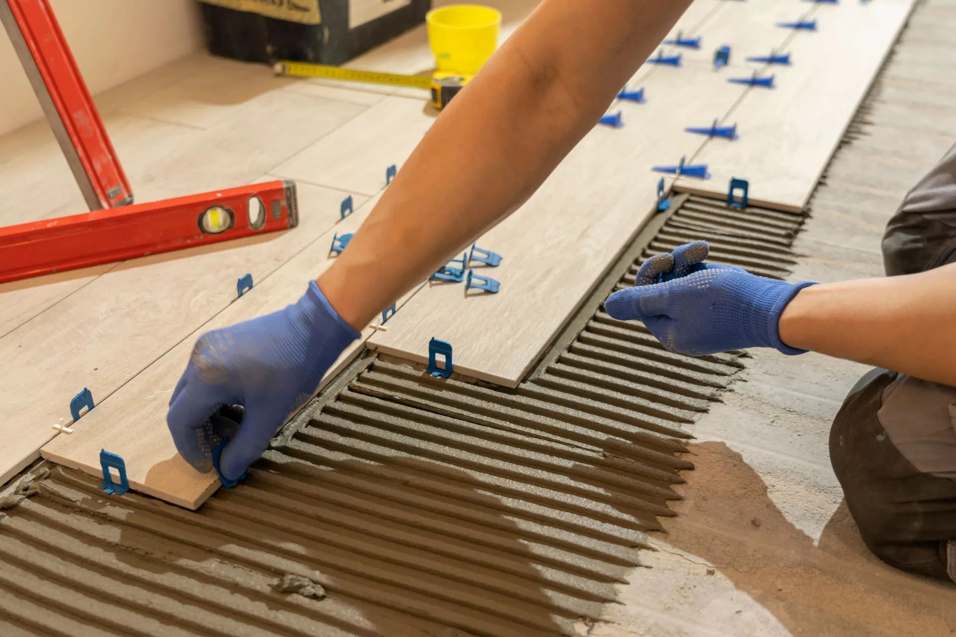 Tile installer spreading thin-set mortar and setting wood-look floor tiles with leveling spacers.