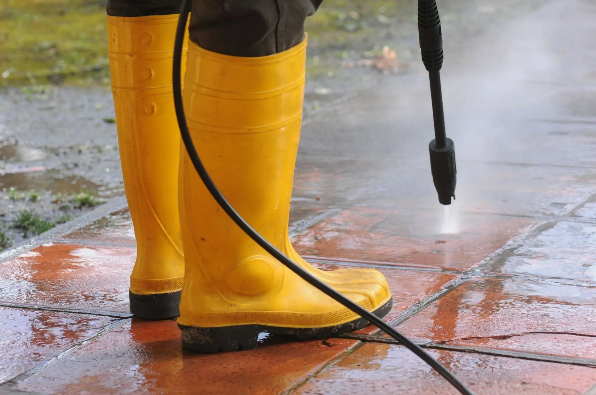 Pressure washer cleaning brick pavers beside yellow work boots during a Troutman, NC driveway and walkway cleaning job